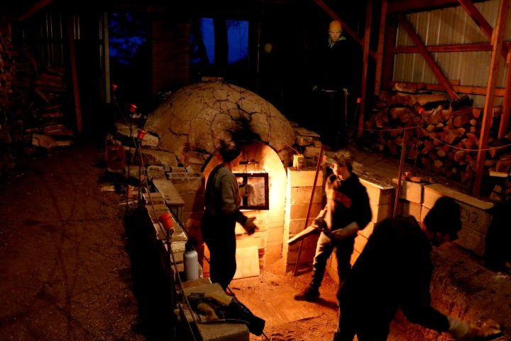 
              People are working around a large wood-fired kiln at night. The kiln is glowing with heat, and a stack of firewood is nearby. The scene is illuminated by the kiln's light and a few lamps, with individuals tending to the fire.